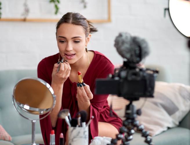 Young woman beauty influencer in red shirt in home setting applying lipstick in front of mirror next to camera mounted on table