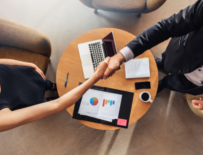 man and woman shake hands across table with laptop and business chart on table