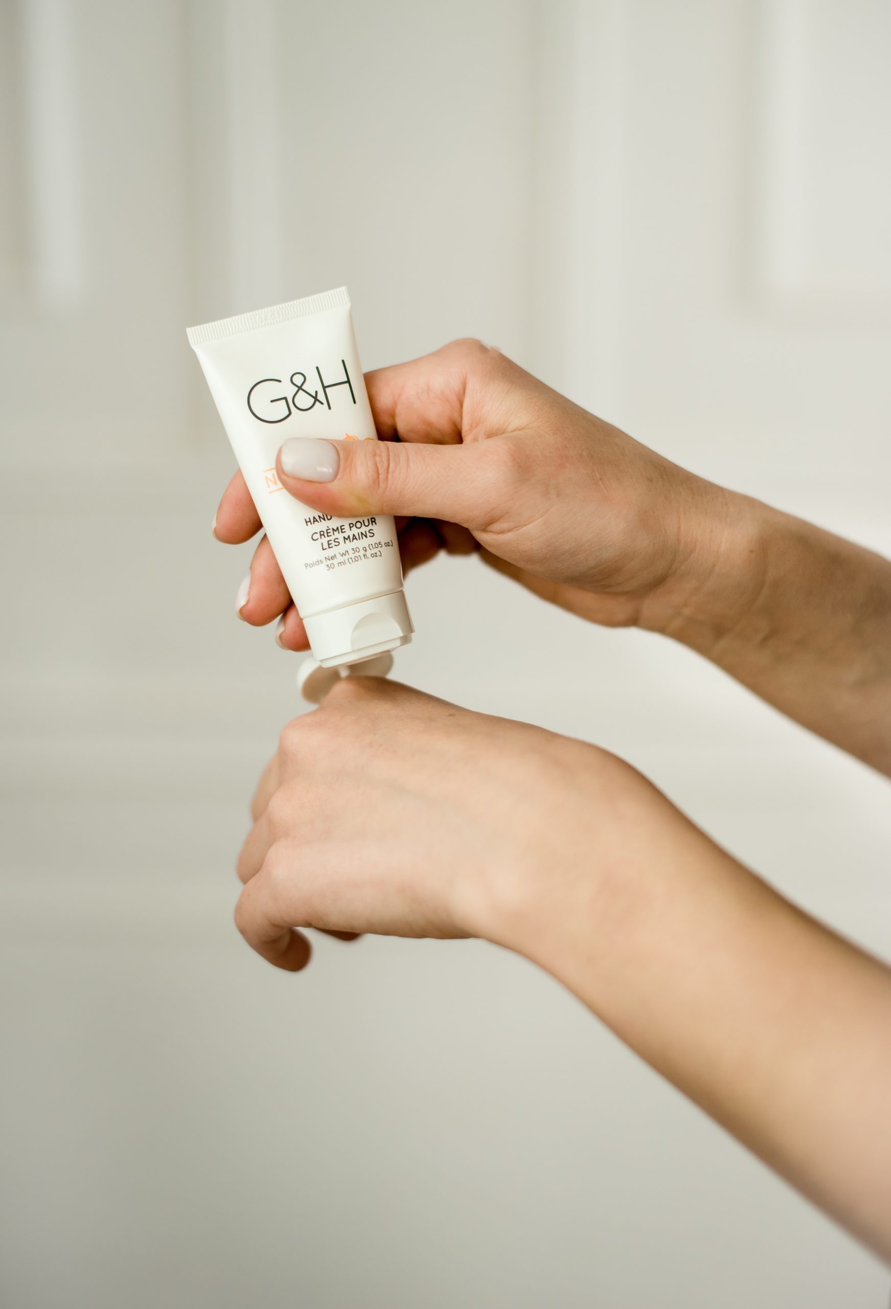 Person applying hand cream from small squeezable container against plain cream background
