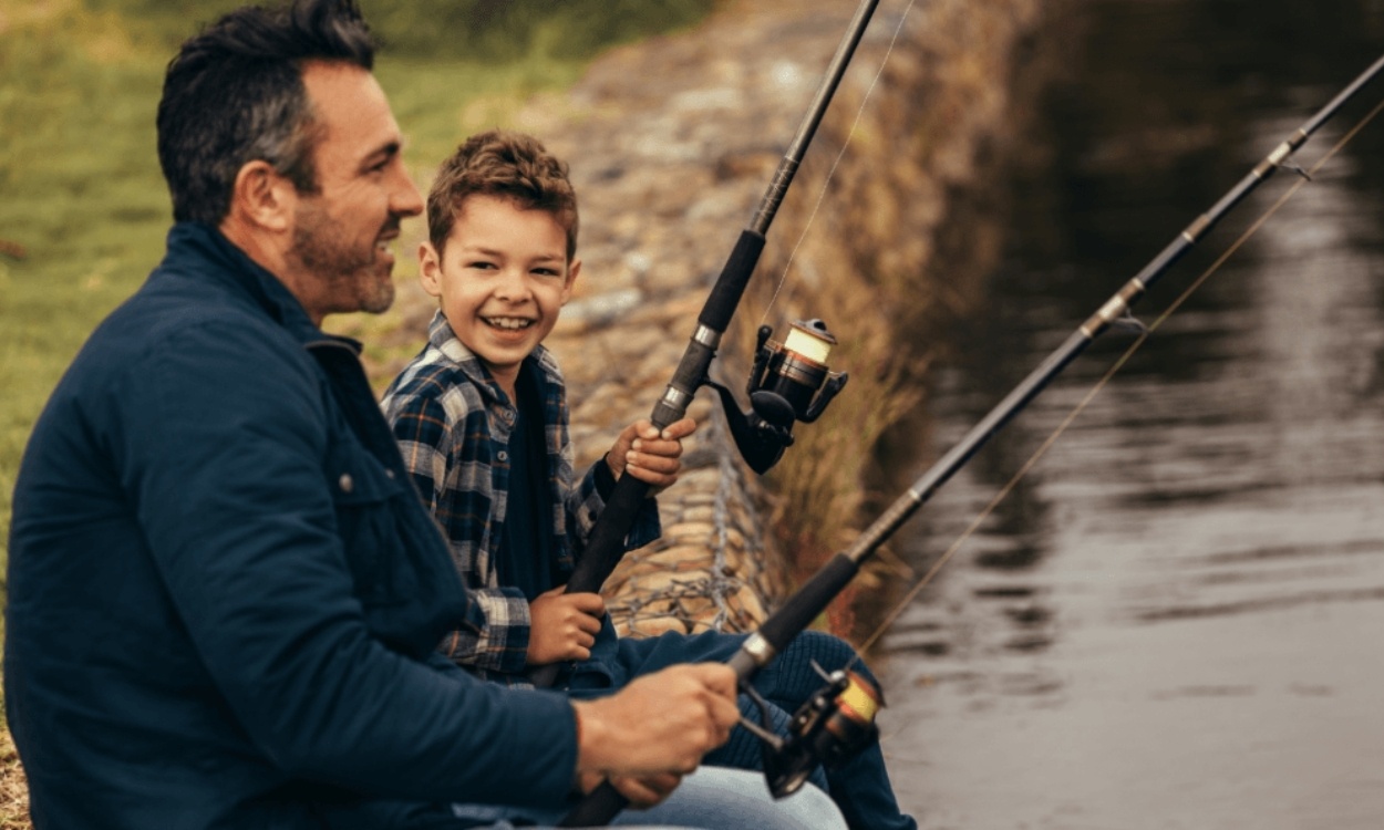 Man and young boy smiling as they fish together, sitting on rocky edge of water