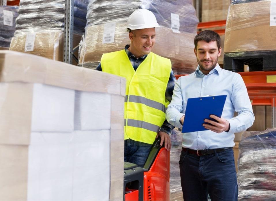 Male sales rep in button down blue shirt in warehouse setting looking at tablet discussing with man in hardhat and industrial wear