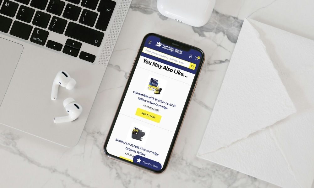 Mobile phone on marble table with CartridgeWorld.co.uk cart page on screen next to laptop and earbuds
