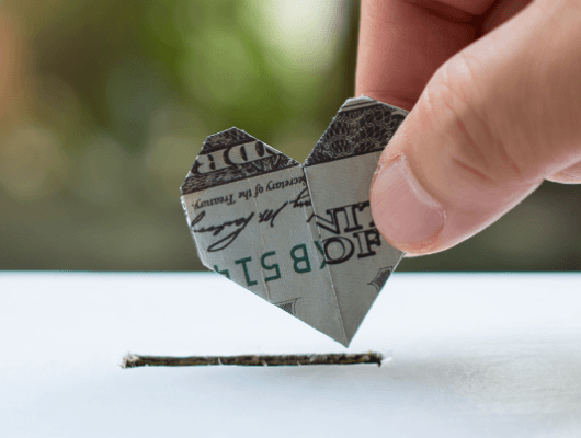 Close-up shot of hand putting heart shaped dollar bill in white box with slit. Charitable giving, caring concept helping to highlight accepting donations for BigCommerce Checkout.
