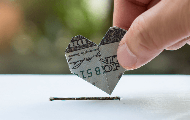 Close-up shot of hand putting heart shaped dollar bill in white box with slit. Charitable giving, caring concept helping to highlight accepting donations for BigCommerce Checkout.