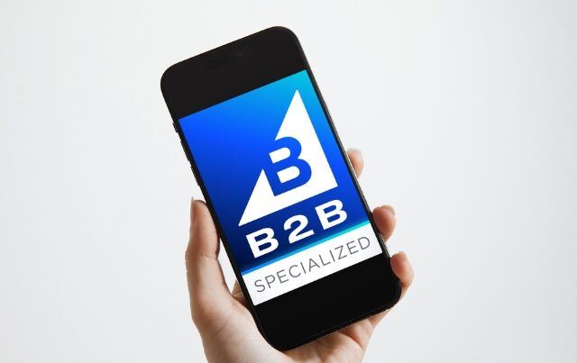 Person holding smartphone against modern gray background with BigCommerce B2B specialized logo on phone screen