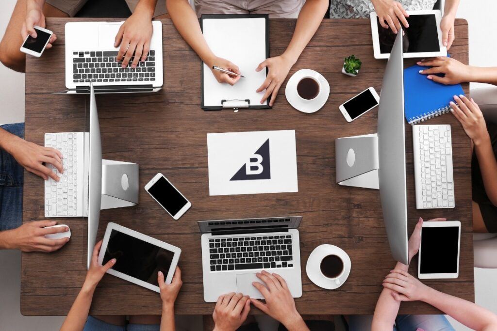 BigCommerce SEO Services - Team working around table with laptops and tablets with BigCommerce logo on paper in the center of table