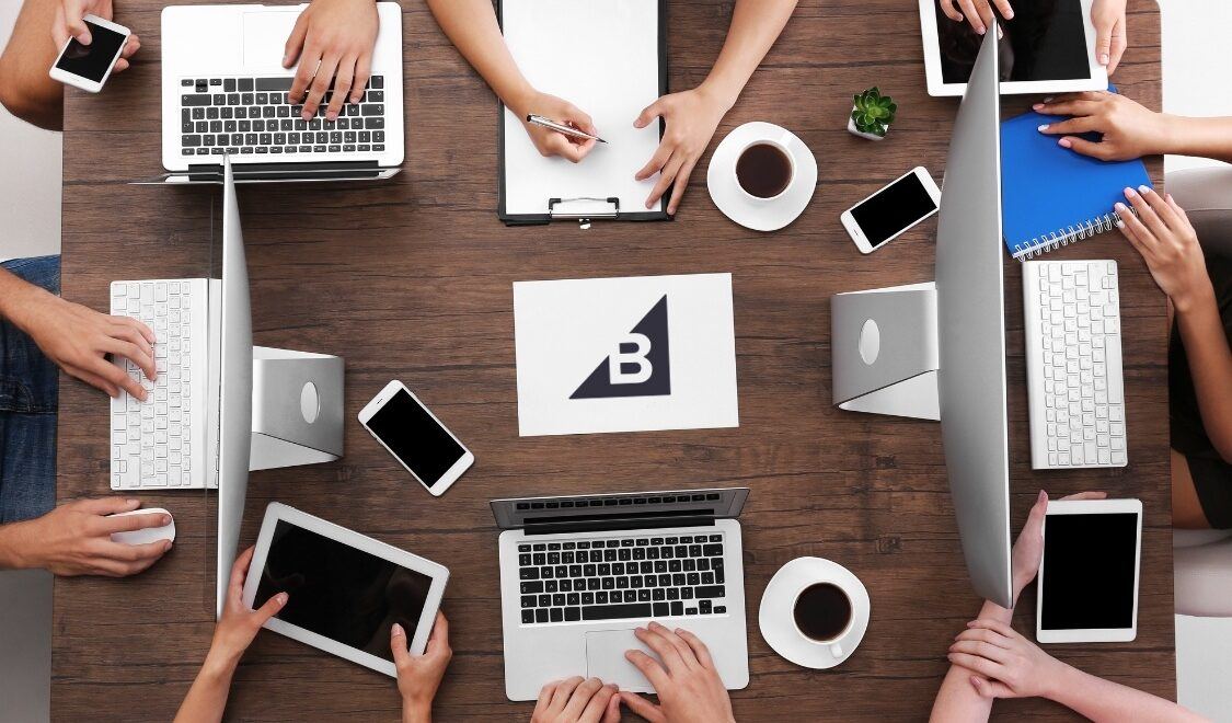BigCommerce SEO Services - Team working around table with laptops and tablets with BigCommerce logo on paper in the center of table