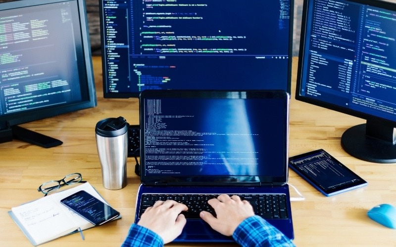 Developer on laptop writing code sitting at desk surrounded by multiple screens - Shopify Technical SEO